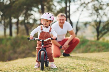 Beyaz gömlekli baba kızına dışarıda bisiklet sürmeyi öğretiyor..