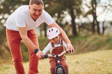 Beyaz gömlekli baba kızına dışarıda bisiklet sürmeyi öğretiyor..