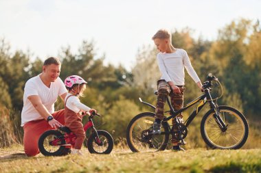 Father with his little daughter and son have fun with bikes outdoors near the forest. clipart