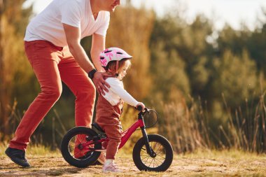 Beautiful sunlight. Father in white shirt teaching daughter how to ride bicycle outdoors. clipart
