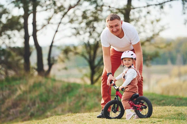 Father in white shirt teaching daughter how to ride bicycle outdoors.