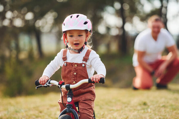 Отец в белой рубашке учит дочь кататься на велосипеде на открытом воздухе.