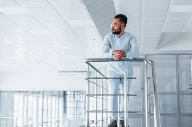 Gümüş renkli korkuluklara yaslanarak. Gündüz vakti ofiste resmi kıyafetler içinde genç yakışıklı bir adam..