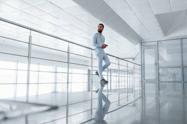 Gümüş renkli korkuluklara yaslanarak. Gündüz vakti ofiste resmi kıyafetler içinde genç yakışıklı bir adam..