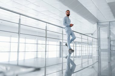 Gümüş renkli korkuluklara yaslanarak. Gündüz vakti ofiste resmi kıyafetler içinde genç yakışıklı bir adam..