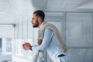Gümüş renkli korkuluklara yaslanarak. Gündüz vakti ofiste resmi kıyafetler içinde genç yakışıklı bir adam..