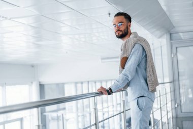 Gümüş renkli korkuluklara yaslanarak. Gündüz vakti ofiste resmi kıyafetler içinde genç yakışıklı bir adam..