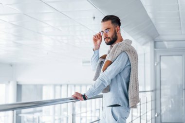 Gümüş renkli korkuluklara yaslanarak. Gündüz vakti ofiste resmi kıyafetler içinde genç yakışıklı bir adam..
