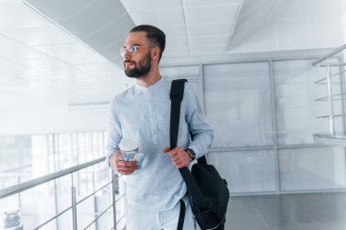 Elinde içki bardağı ve siyah çanta var. Gündüz vakti ofiste resmi kıyafetler içinde genç yakışıklı bir adam..