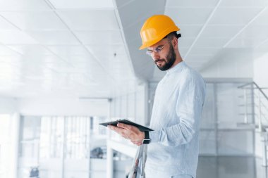 Holding notepad. Engineer in white clothes and orange protective hard hat standing and working indoors.