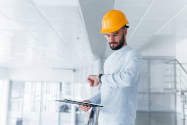 Holding notepad. Engineer in white clothes and orange protective hard hat standing and working indoors.