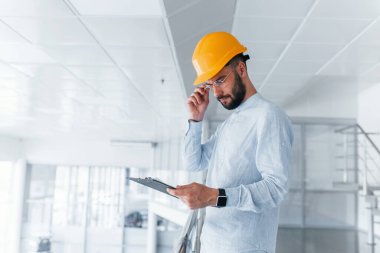 Holding notepad. Engineer in white clothes and orange protective hard hat standing and working indoors.