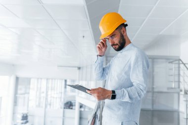 Holding notepad. Engineer in white clothes and orange protective hard hat standing and working indoors.