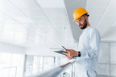 Holding notepad. Engineer in white clothes and orange protective hard hat standing and working indoors.
