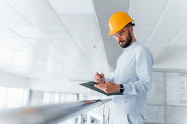 Holding notepad. Engineer in white clothes and orange protective hard hat standing and working indoors.