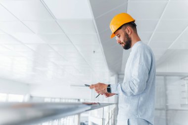 Holding notepad. Engineer in white clothes and orange protective hard hat standing and working indoors.