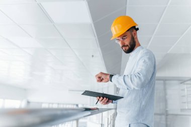 Holding notepad. Engineer in white clothes and orange protective hard hat standing and working indoors.