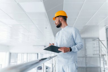 Holding notepad. Engineer in white clothes and orange protective hard hat standing and working indoors.
