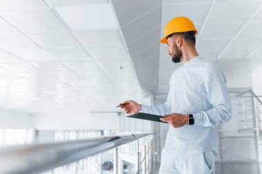 Holding notepad. Engineer in white clothes and orange protective hard hat standing and working indoors.