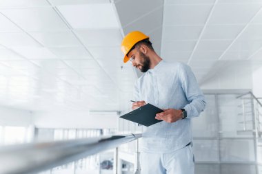 Holding notepad. Engineer in white clothes and orange protective hard hat standing and working indoors.