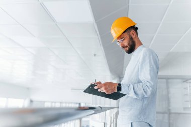 Holding notepad. Engineer in white clothes and orange protective hard hat standing and working indoors.