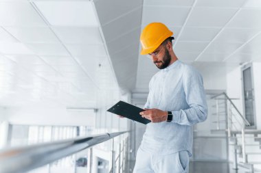 Holding notepad. Engineer in white clothes and orange protective hard hat standing and working indoors.