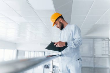 Holding notepad. Engineer in white clothes and orange protective hard hat standing and working indoors.