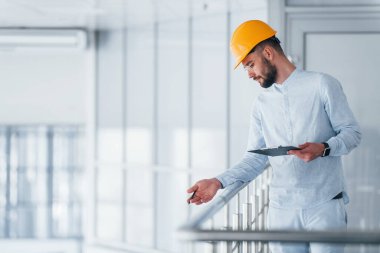 Holding notepad. Engineer in white clothes and orange protective hard hat standing and working indoors.