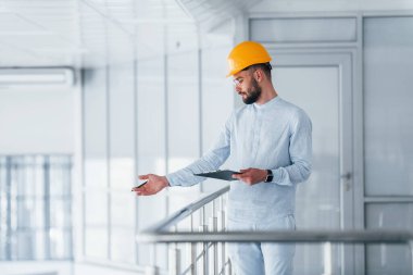 Not defterini tutuyorum. Beyaz elbiseli ve turuncu koruyucu şapkalı mühendis içeride duruyor ve çalışıyor..