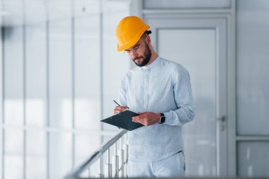 Not defterini tutuyorum. Beyaz elbiseli ve turuncu koruyucu şapkalı mühendis içeride duruyor ve çalışıyor..