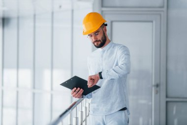 Not defterini tutuyorum. Beyaz elbiseli ve turuncu koruyucu şapkalı mühendis içeride duruyor ve çalışıyor..