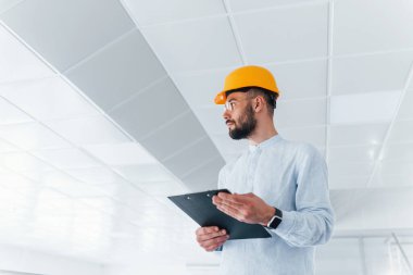 Beyaz giysili ve turuncu şapkalı mühendis ön planda. Kapalı alanda ayakta ve çalışıyor..