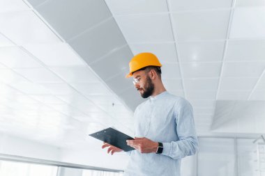 Front view of engineer in white clothes and orange protective hard hat that standing and working indoors.