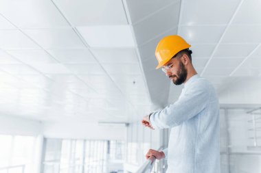Front view of engineer in white clothes and orange protective hard hat that standing and working indoors.