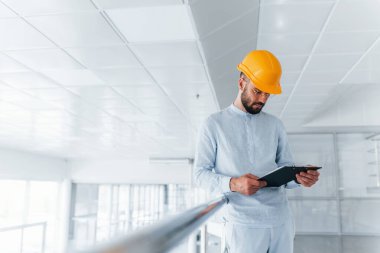 Beyaz giysili ve turuncu şapkalı mühendis ön planda. Kapalı alanda ayakta ve çalışıyor..