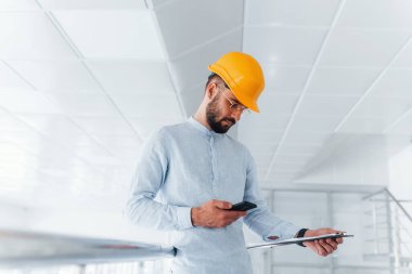 Using phone. Engineer in white clothes and orange protective hard hat standing and working indoors.
