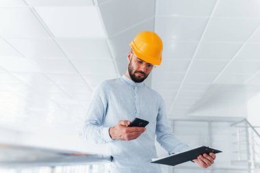 Telefonla. Beyaz elbiseli ve turuncu koruyucu şapkalı mühendis içeride duruyor ve çalışıyor..