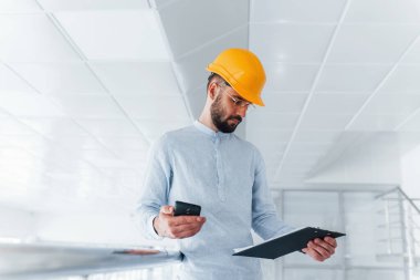 Using phone. Engineer in white clothes and orange protective hard hat standing and working indoors.
