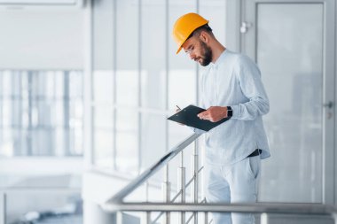 Walking near silver colored railings. Engineer in white clothes and orange protective hard hat standing and working indoors.