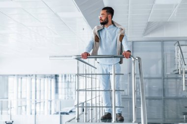 Gümüş renkli korkuluklara yaslanarak. Gündüz vakti ofiste resmi kıyafetler içinde genç yakışıklı bir adam..