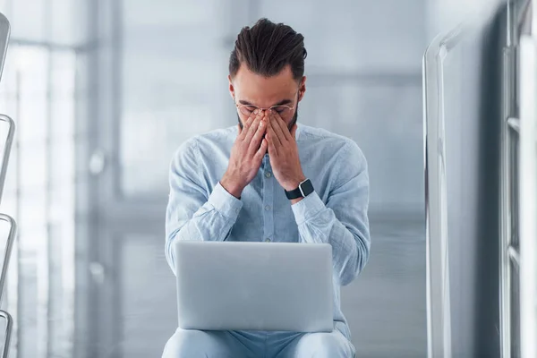 Feeling tired and bad. Sitting on the stairs with laptop. Young handsome man in formal clothes indoors in the office at daytime.