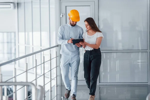 Talking with young girl in casual clothes. Engineer in white clothes and orange protective hard hat standing and working indoors.