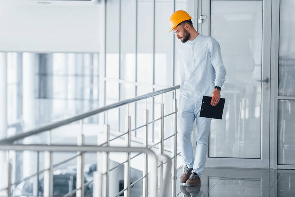 Walking near silver colored railings. Engineer in white clothes and orange protective hard hat standing and working indoors.