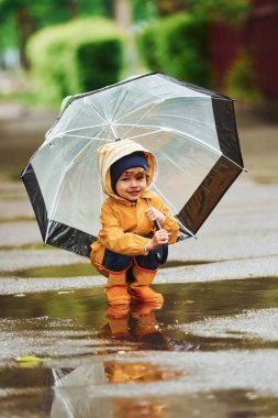 Kid in yellow waterproof cloak, boots and with umbrella playing outdoors after the rain.