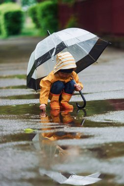 Kid in yellow waterproof cloak, boots and with umbrella playing outdoors after the rain.