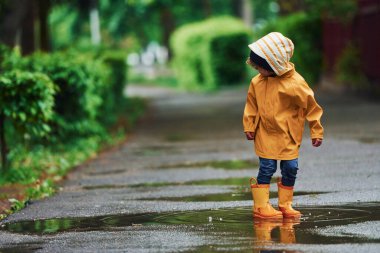 Kid in yellow waterproof cloak and boots playing outdoors after the rain.