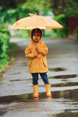 Kid in yellow waterproof cloak, boots and with umbrella playing outdoors after the rain.