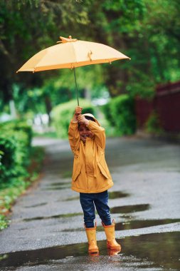 Kid in yellow waterproof cloak, boots and with umbrella playing outdoors after the rain.