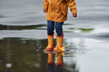 Sarı su geçirmez pelerinli ve botlu çocuk yağmurdan sonra el yapımı tekne oyuncağıyla oynuyor..