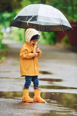 Kid in yellow waterproof cloak, boots and with umbrella playing outdoors after the rain.